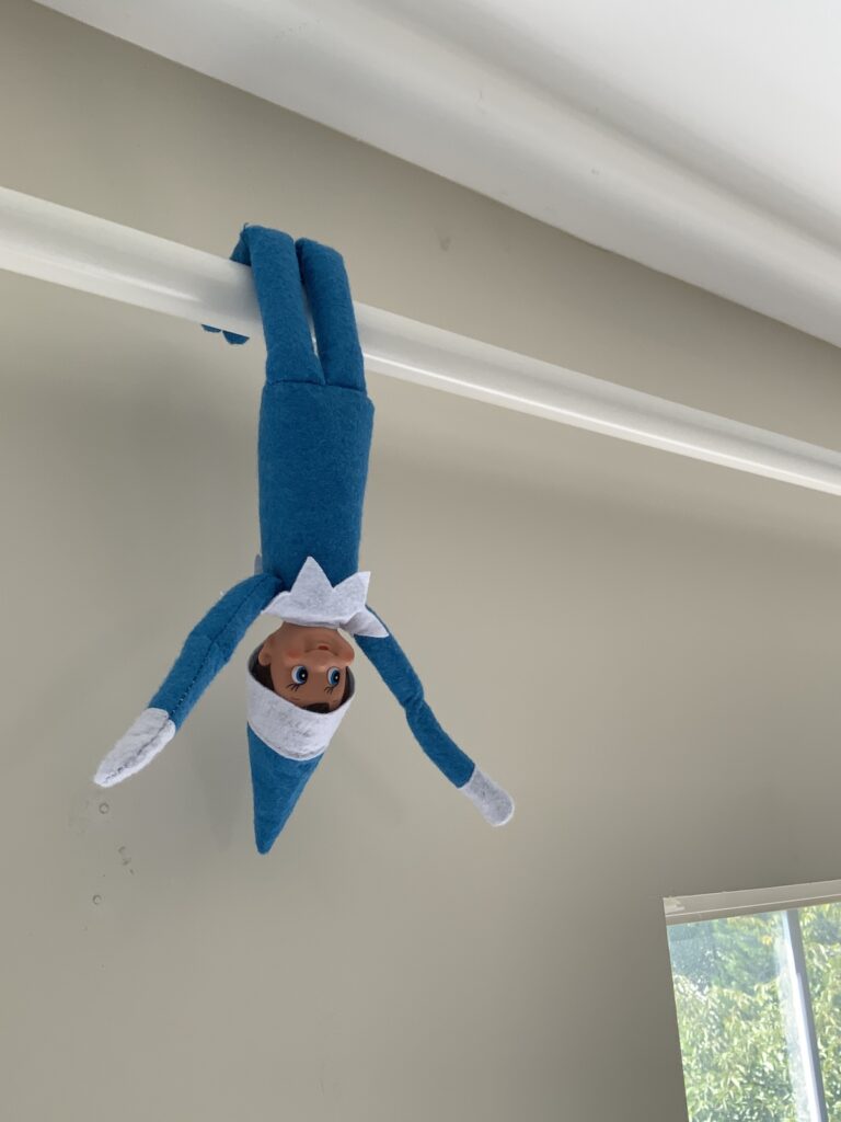 An elf on the shelf doll hanging over a curtain rod by its legs.