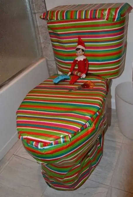 An elf on the shelf doll sitting on a toilet that has been wrapped in Christmas-themed wrapping paper.