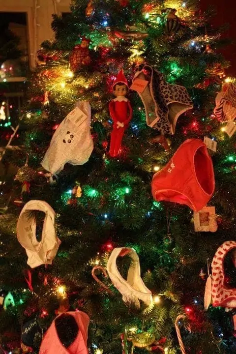 An elf on the shelf doll in a Christmas tree with underwear in the tree like decorations.