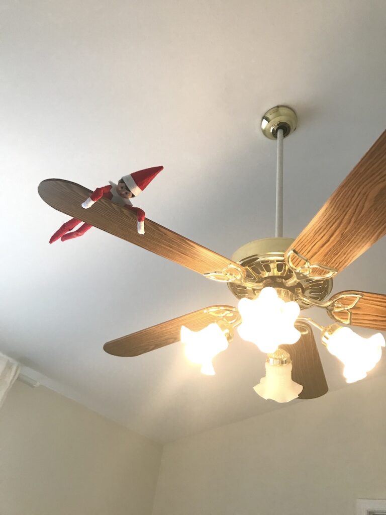 An elf on the shelf doll holding on to the ceiling fan blade.