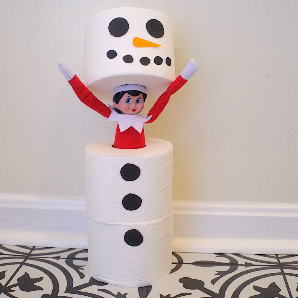 An elf on the shelf popping out of the center of a snowman made of three rolls of toilet paper.