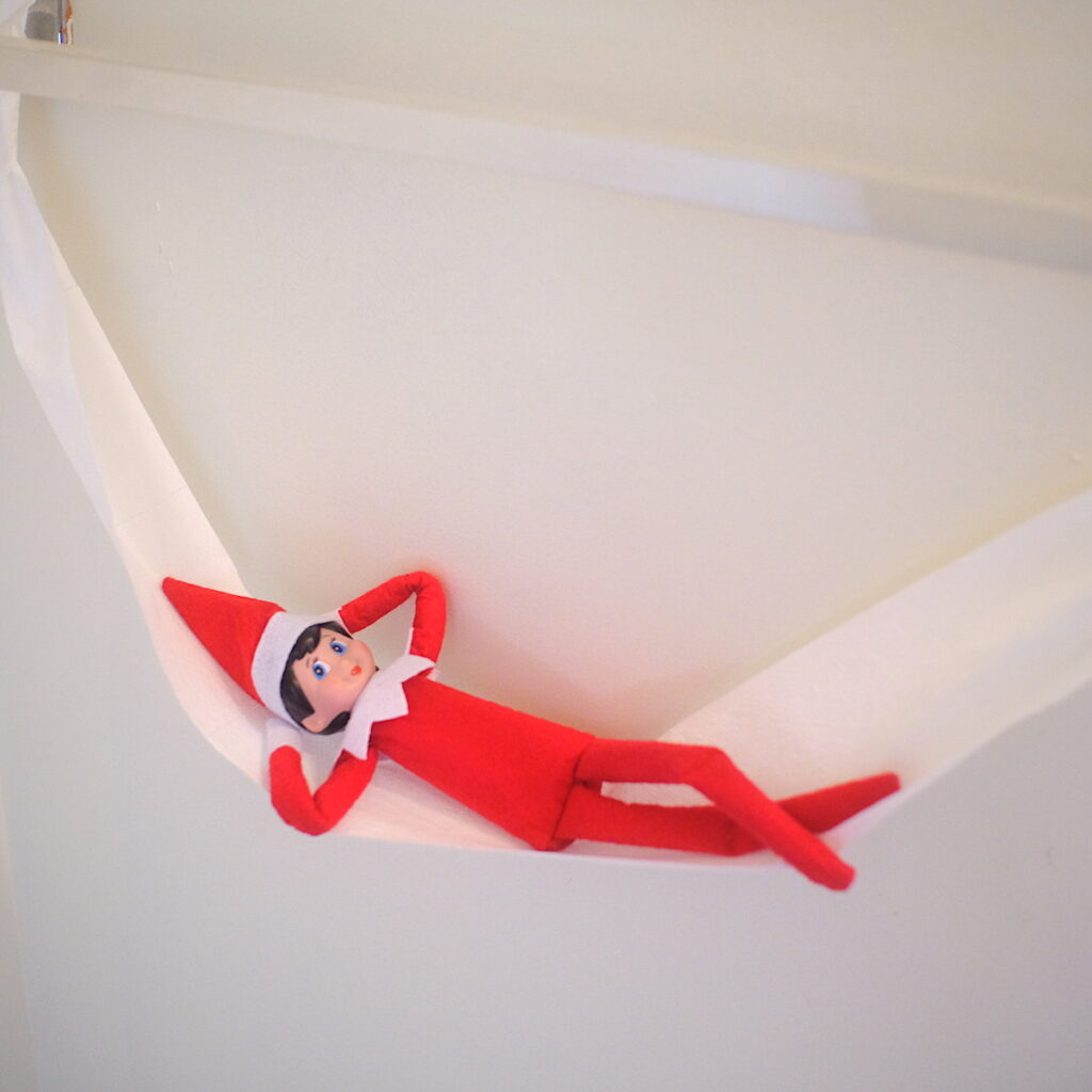 An elf on the shelf doll laying comfortably in a hammock made of toilet paper.