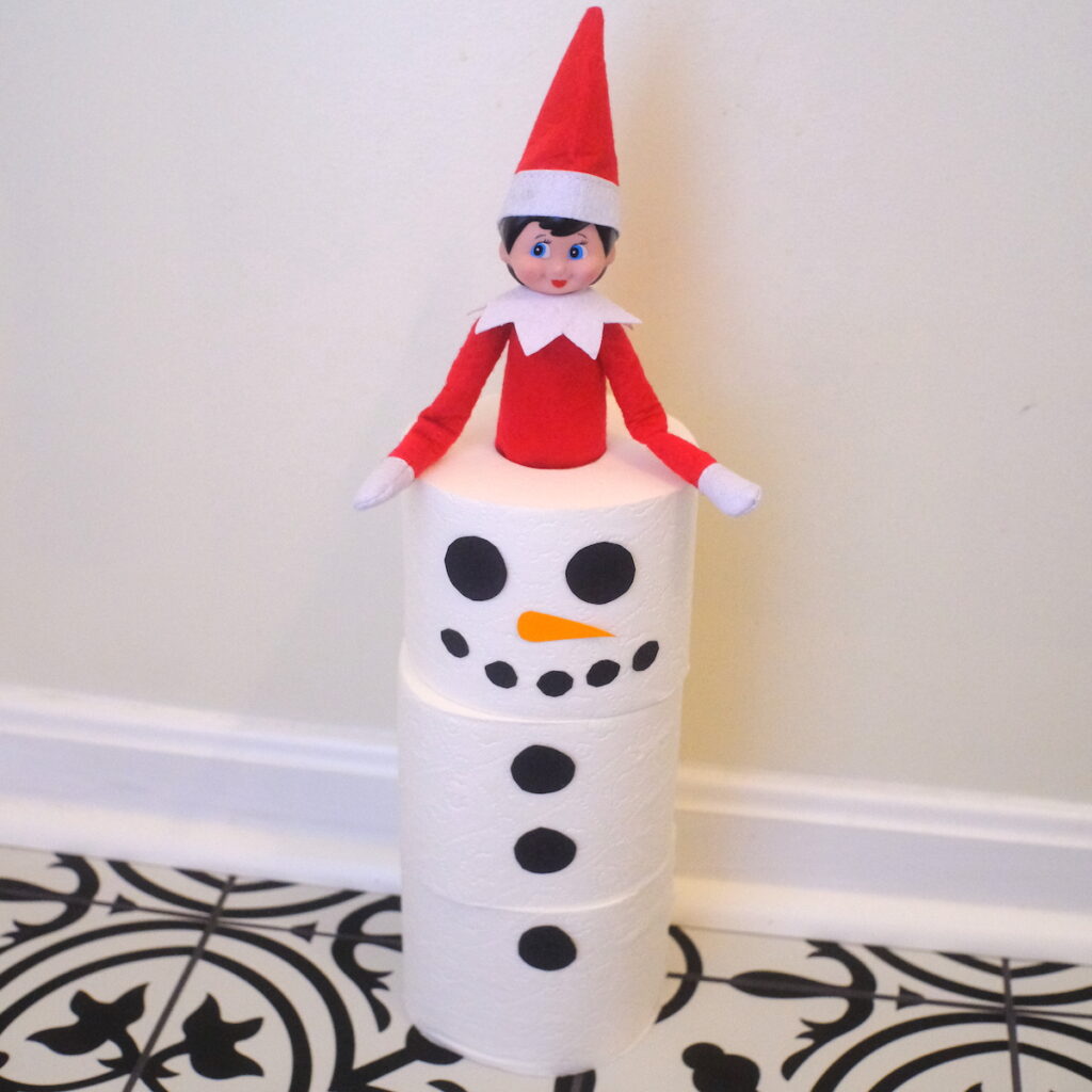 An elf on the shelf popping out of the top of a snowman made of three rolls of toilet paper.