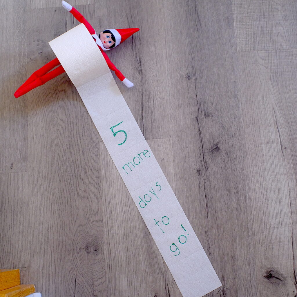 An elf on the shelf doll inside a roll of toilet paper with the words "5 more days to go" written on the paper.