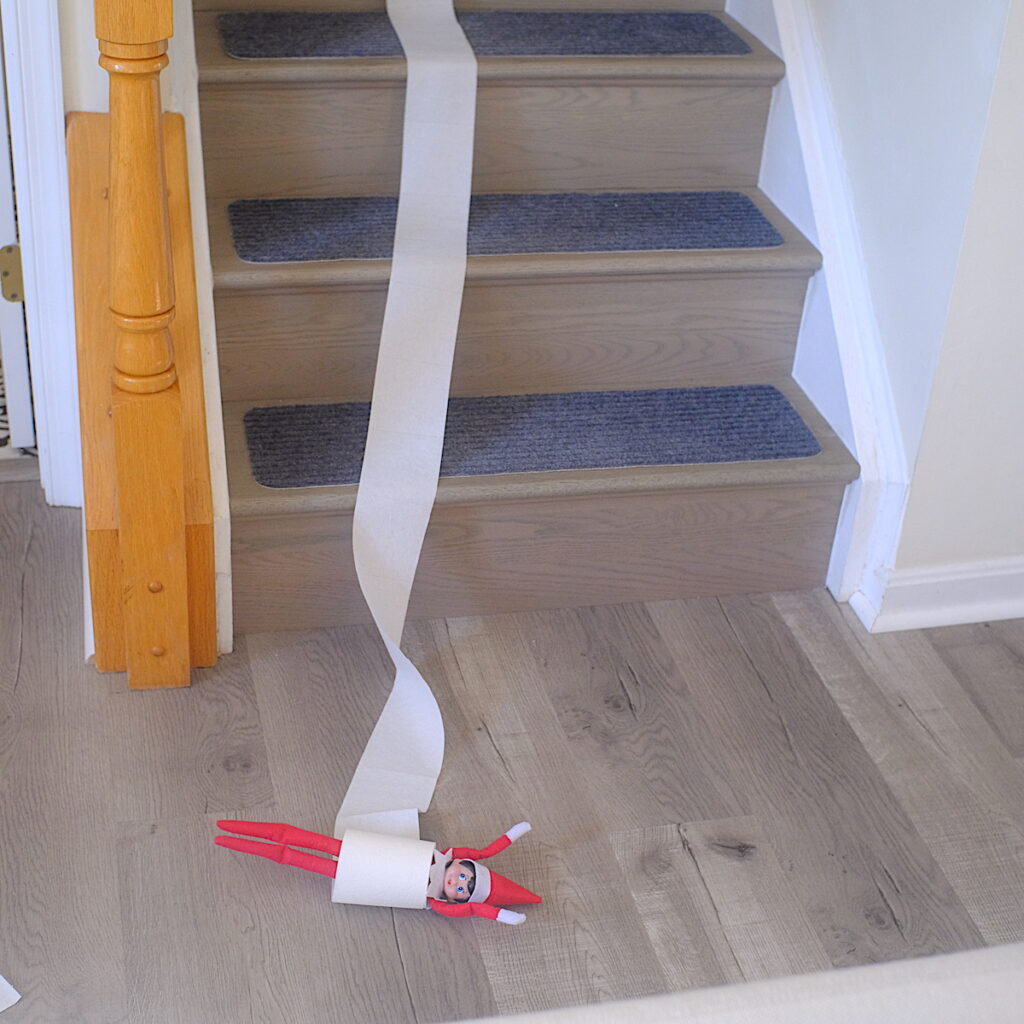 An elf on the shelf rolling down the stairs in the center of a roll of toilet paper.