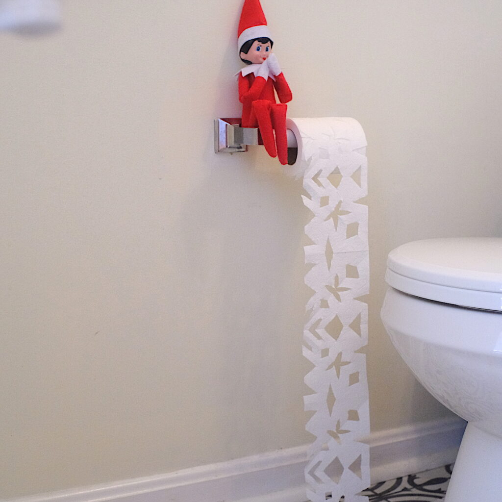 An elf on the shelf doll sitting next to a roll of toilet paper that has been cut to look like paper snowflakes.