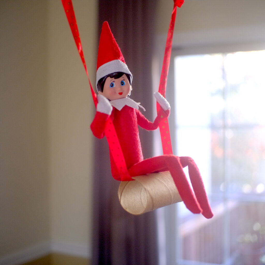An elf on the shelf doll swinging on a toilet paper tube.