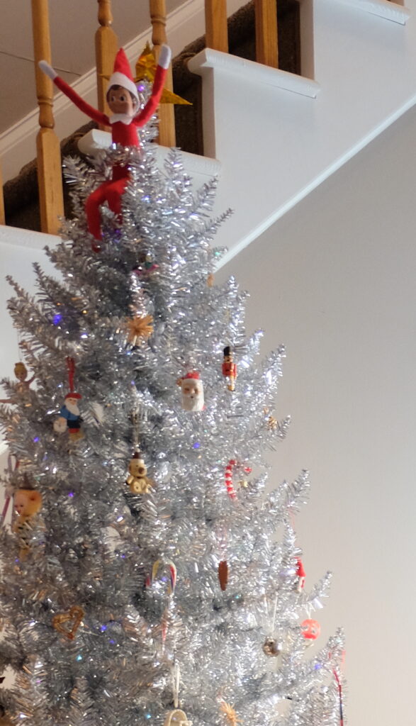 an elf on the shelf doll perched in a silver christmas tree