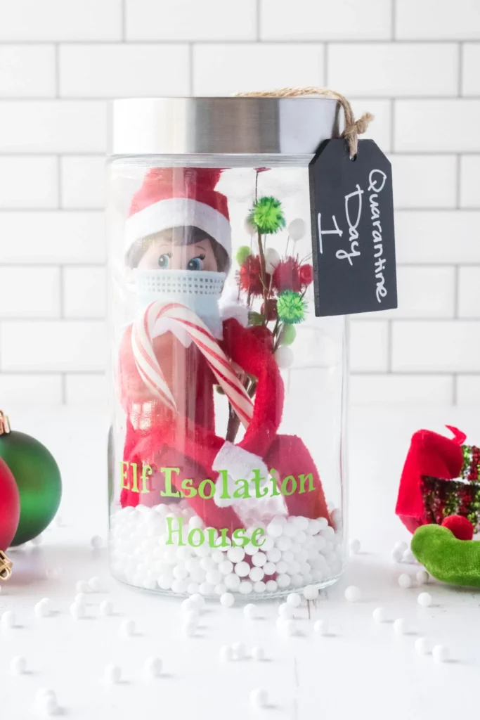 an elf on the shelf doll inside a glass container, wearing a face mask and the jar is labeled "Elf Isolation Jar, Quarantine Day 1"