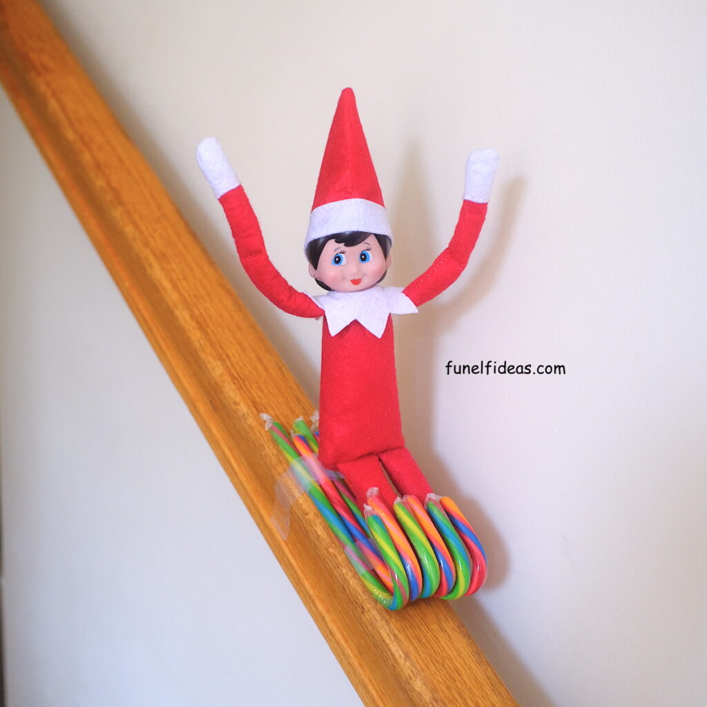 A girl elf on the shelf doll sliding down a candy cane sled that is taped to the stairs railing.