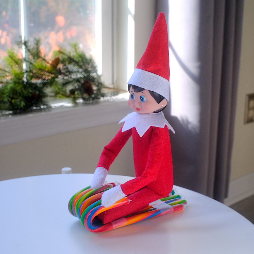 A girl elf on the shelf doll sitting in a sled made by taping candy canes together.