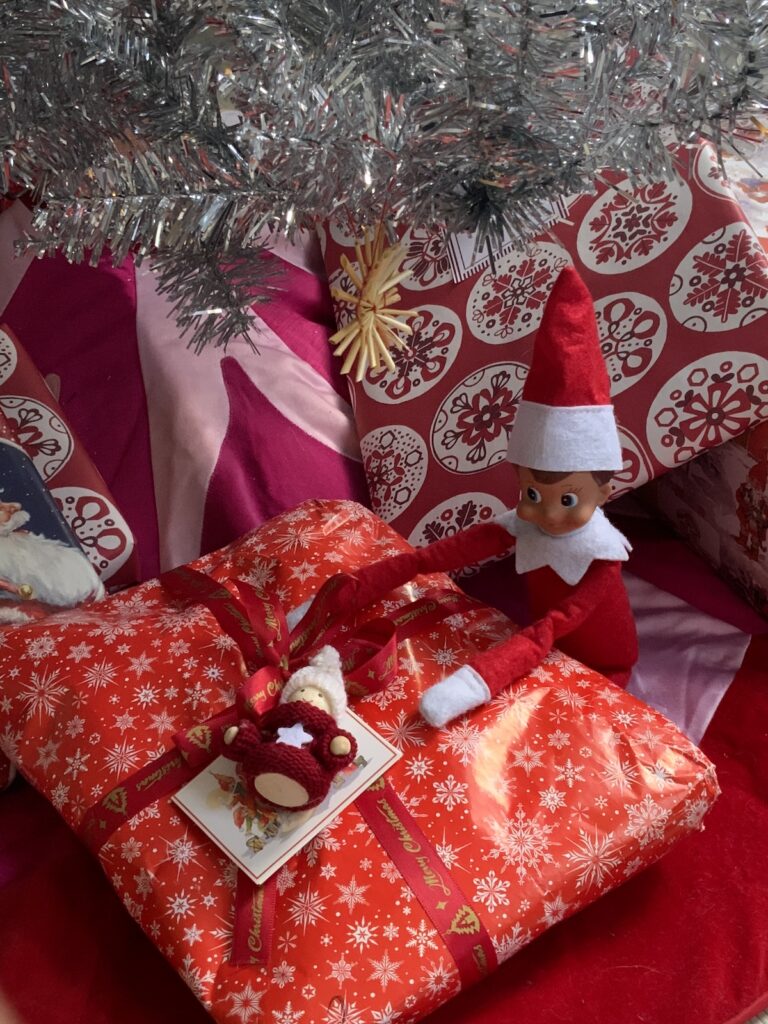 An elf on the shelf doll sitting in the Christmas presents under a silver christmas tree.