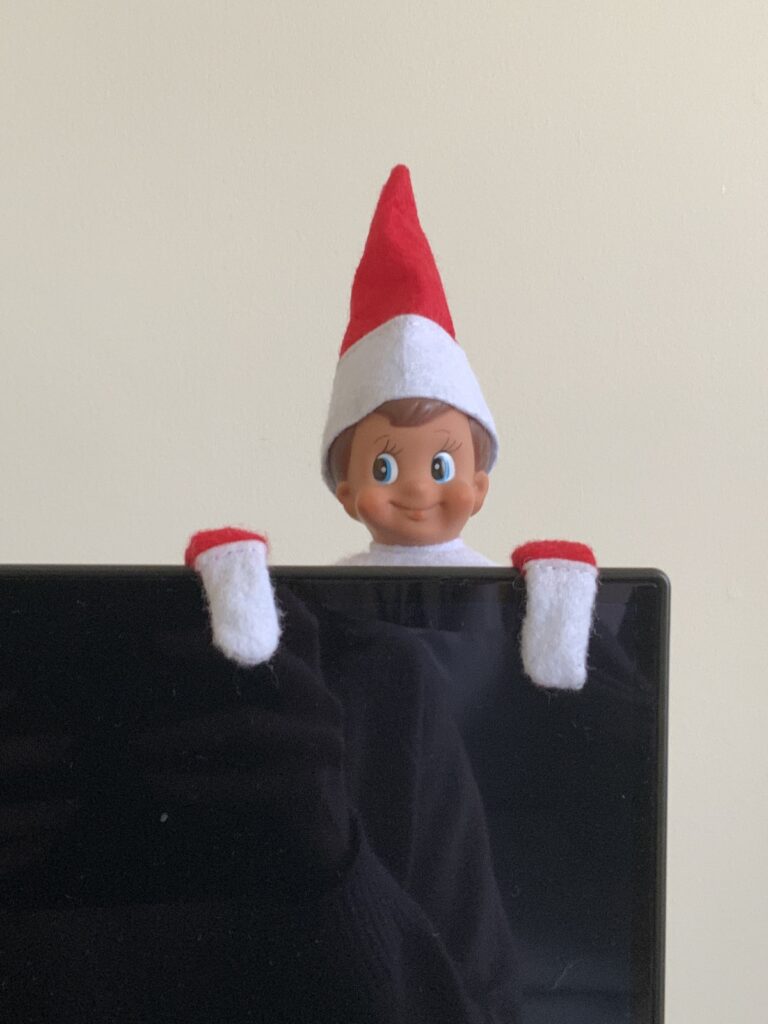 a boy elf on the shelf doll peeking over the top of a TV.