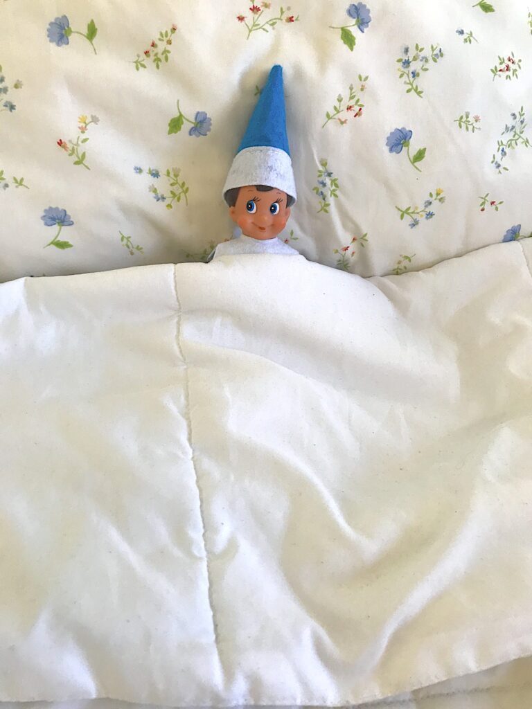 An elf on the shelf doll tucked into the covers on a bed.
