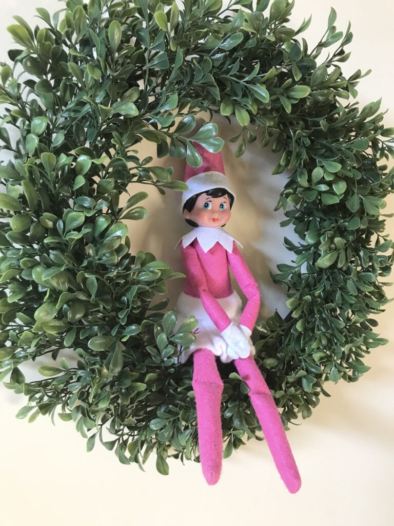 An elf on the shelf doll sitting in a wreath.