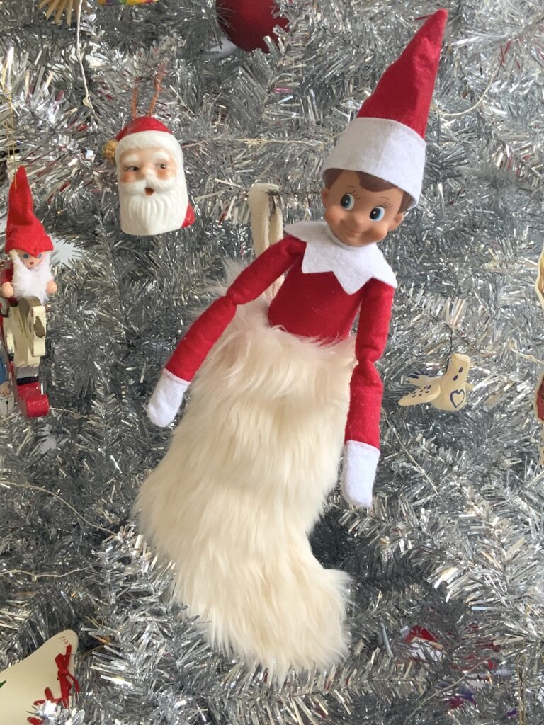 a boy elf on the shelf doll with a red outfit peeking out of a white furry christmas stocking.