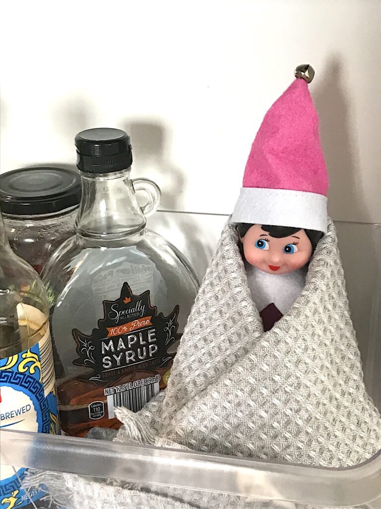 An elf on the shelf doll wrapped in a towel sitting in the fridge door shelf.