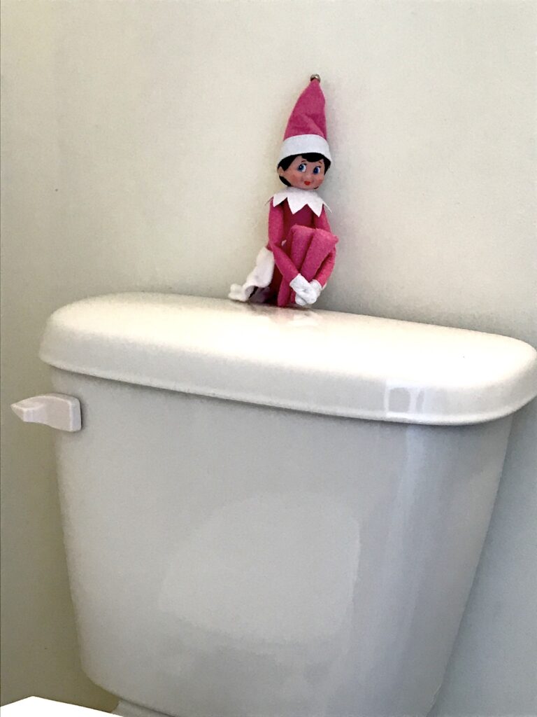 An elf on the shelf doll sitting on top of a toilet.