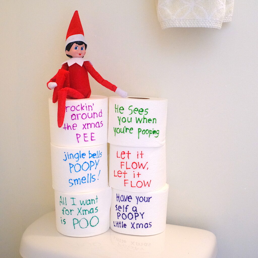 An elf on the shelf doll sitting on top of 6 rolls of toilet paper that have funny potty humor written on them.