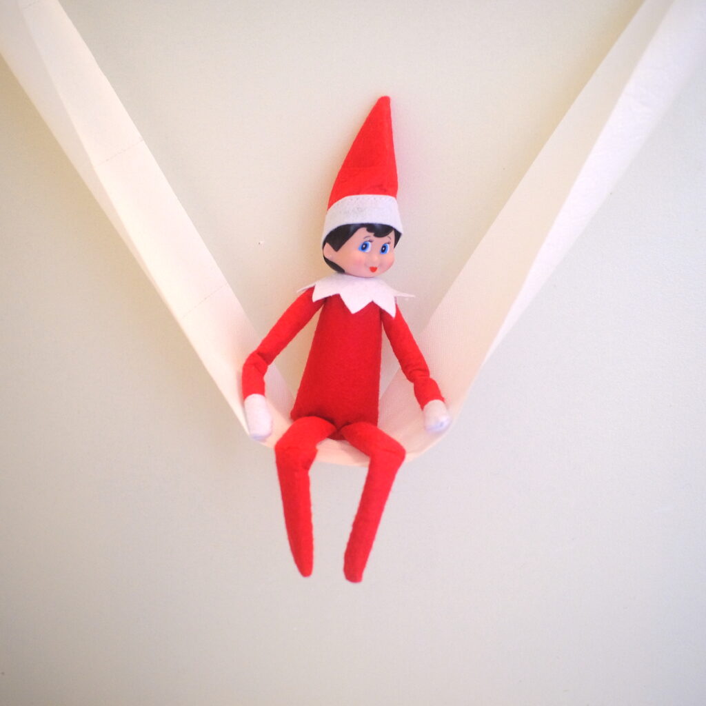 An elf on the shelf doll sitting on a swing made of toilet paper.