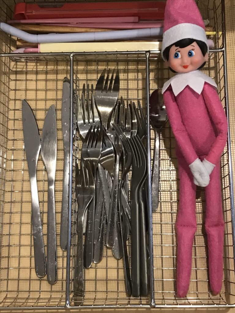 An elf on the shelf doll laying in a kitchen utensil organizer in the drawer.