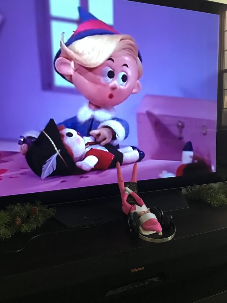 An elf on the shelf doll watching a christmas movie on tv while holding a remote.